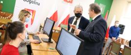 Wojewoda opolski odwiedza punkt rejestracji nr pesel w UG Gogolin. Na zdjęciu po lewej stronie dwie pracownice urzędu, siedzące przy biurku. Przed nimi komputer. Na przeciw nich, stoją wojewoda opolski i burmistrz gminy. Spoglądają na siebie, rozmawiają. W tle biało-czerwone flagi, stół oraz pozostali uczestnicy spotkania