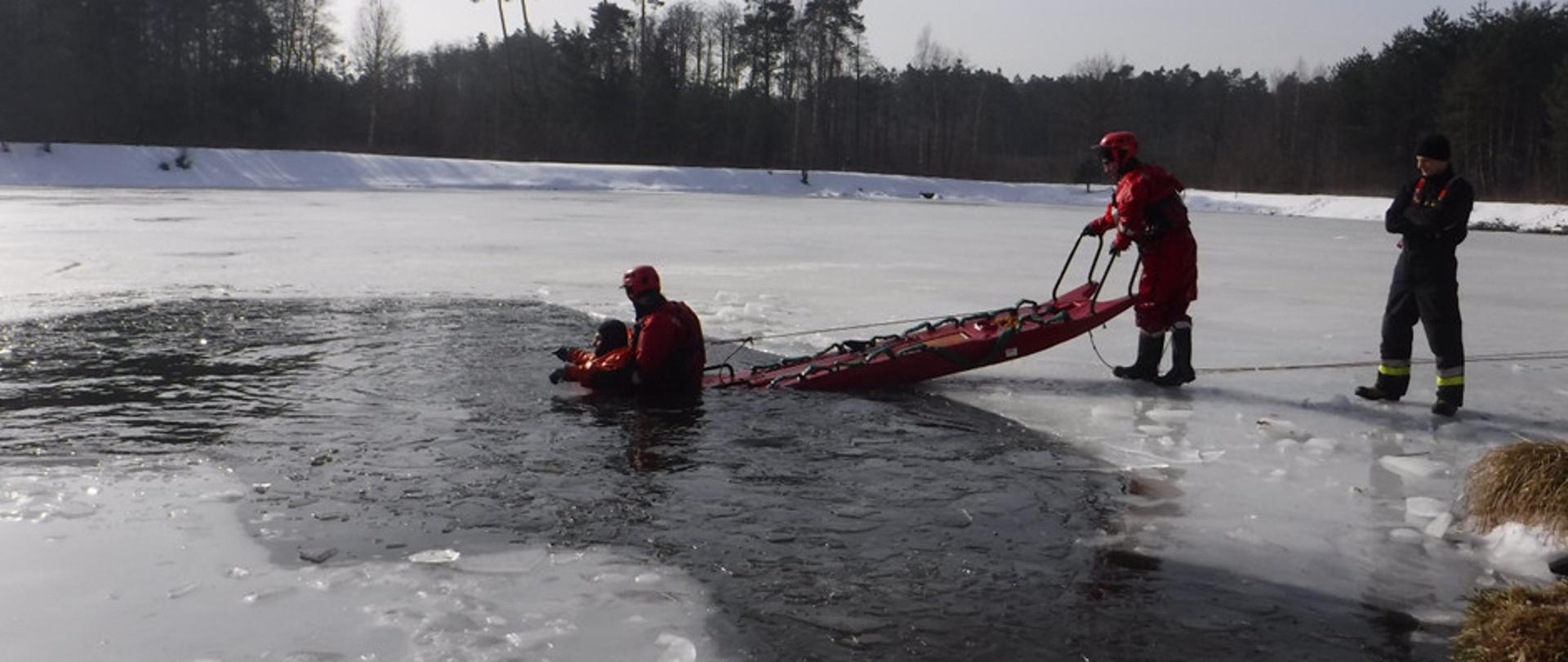Na zdjęciu widzimy zbiornik wodny pokryty lodem z wybitą przeręblą. W przerębli znajduję się pozorant ubrany w sprzęt specjalistyczny, którego zabezpiecza ratownik w ubraniu specjalistycznym odpowiednio zabezpieczony, kolejny ratownik operuje saniami lodowymi. Ratownicy podejmują czynności służące wyciągnięciu pozoranta z przerębli. Wszystko nadzoruje stojący po lewej stronie zdjęcia dowódca.