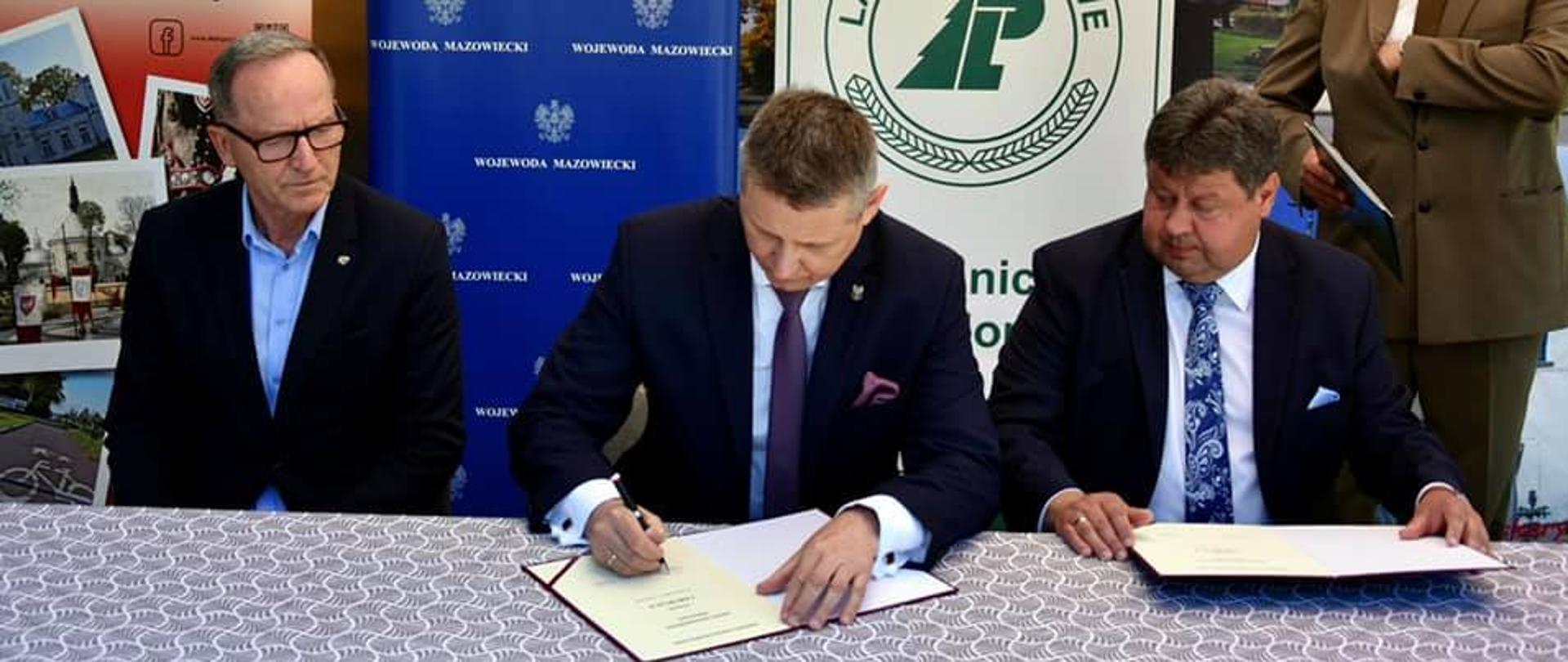 Zdjęcie przedstawia moment podpisania umowy na dofinansowanie zadania w ramach Rządowego Funduszu Rozwoju Dróg. Na zdjęciu znajduje się trzech elegancko ubranych mężczyzn, którzy siedzą za stołem. Dwóch mężczyzn jest w trakcie podpisywania umowy. 