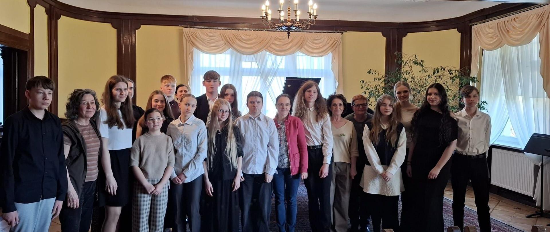 Zdjęcie kolorowe. Grupa kilkunastu osób ustawiona do pamiątkowej fotografii w eleganckim, klasycznym wnętrzu. Osoby ubrane są głównie w stroje galowe: białe koszule, ciemne spodnie, spódnice oraz czarne bluzki. W centralnej części pomieszczenia wisi żyrandol z zapalonymi żarówkami imitującymi płomienie świec. W tle znajdują się okna z jasnymi, upiętymi firanami, a nad nimi drewniana boazeria z godłem Polski (orzeł w koronie na czerwonej tarczy). Po prawej stronie stoi duża roślina doniczkowa oraz fragment pianina. Podłoga wykonana z jasnego parkietu, częściowo przykryta wzorzystym dywanem. Na pierwszym planie widoczne są oparcia trzech drewnianych krzeseł. Ściany mają żółty odcień i są przedzielone pionowymi, ciemnymi elementami konstrukcyjnymi. Wszystkie osoby stoją blisko siebie, patrząc w stronę obiektywu.