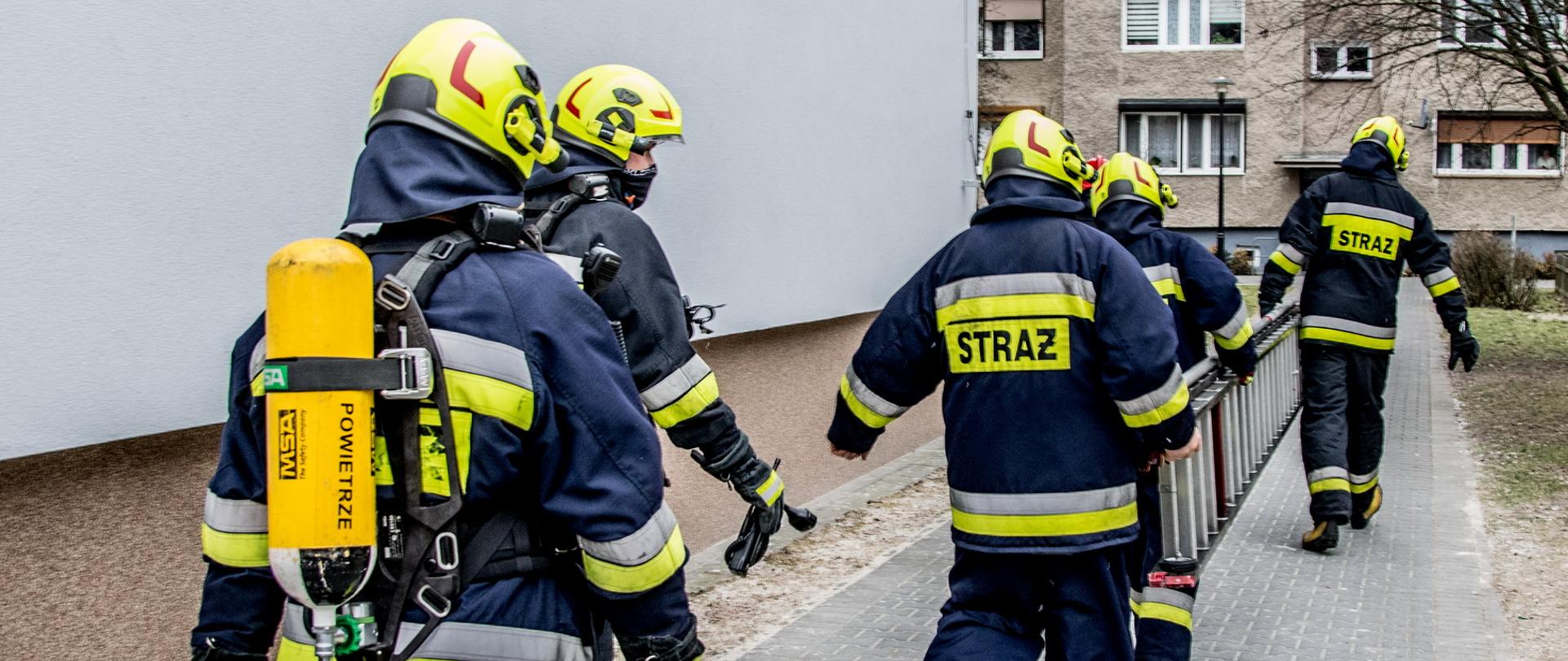 Strażacy niosą drabinę. Dwóch z nich ma ubrane aparaty powietrzne.