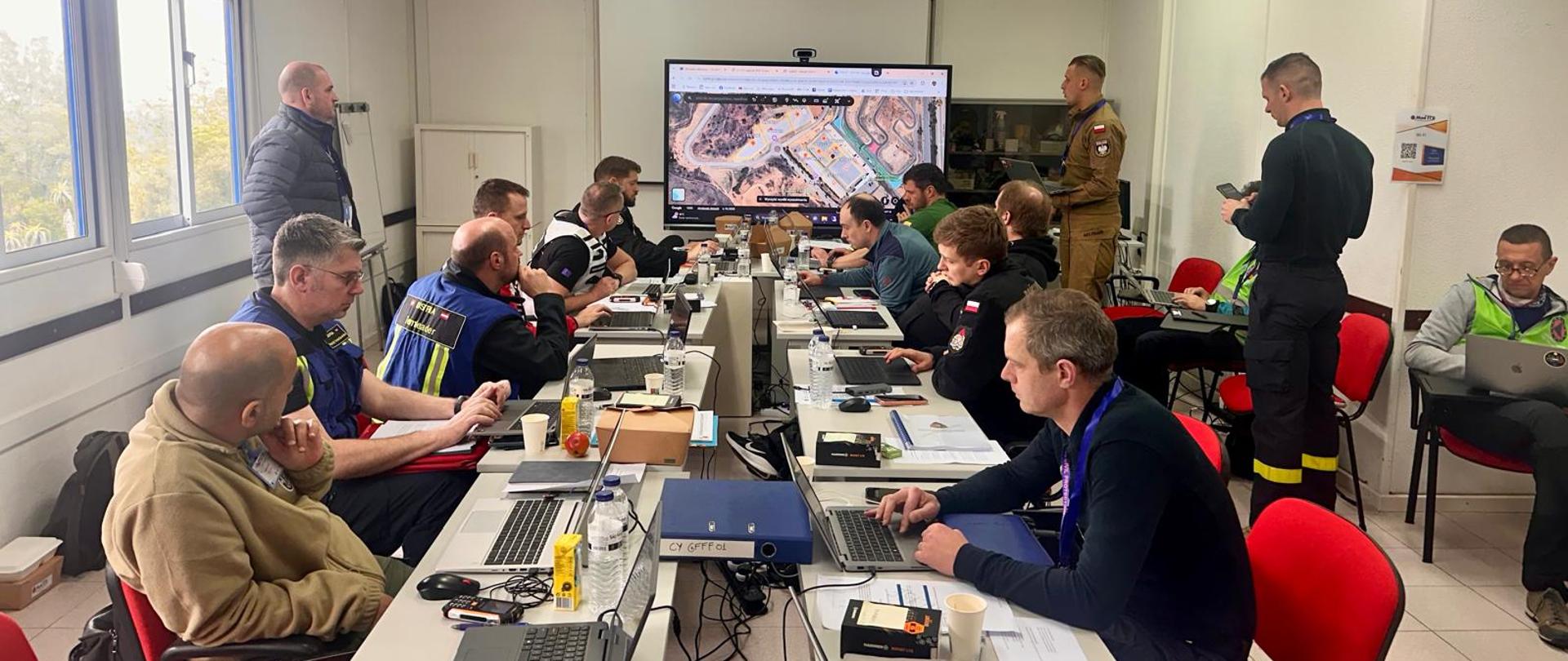 Na zdjęciu uczestnicy ćwiczeń zajmują miejsca za stołami, reprezentując różne podmioty ratownicze - są ubrani w kamizelki określające funkcje i podmiot. Przed nimi znajduje się osoba kierująca ćwiczeniami, również ubrana w kamizelkę. Wszyscy znajdują się wewnątrz sali konferencyjnej. W tle widać ekran do prezentowania materiału elektronicznego. 
