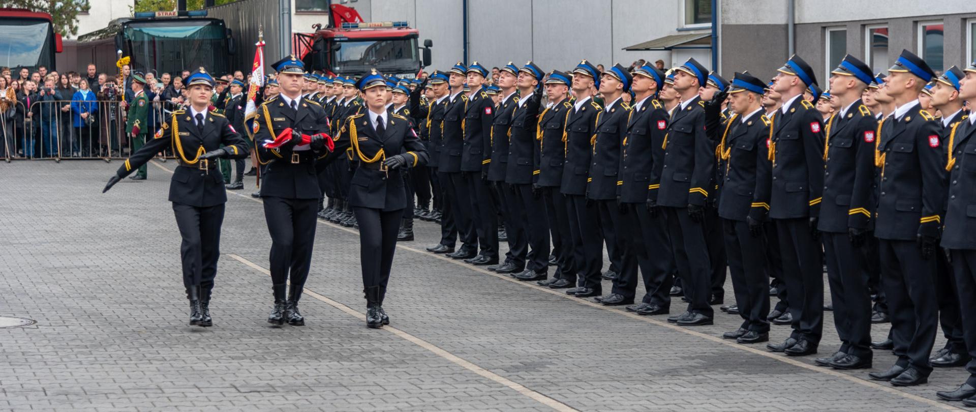 Trzy strażaczki PSP w galowych mundurach wnoszą flagę państwową podczas uroczystości ślubowania strażaków.
