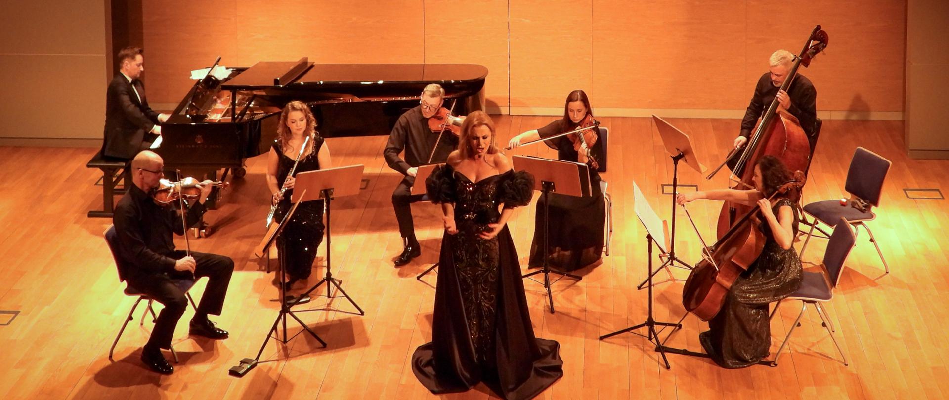 Szeroki kadr prezentujący cały zespół na scenie z perspektywy lotu ptaka. Na środku stoi solistka, otoczona przez siedmioosobowy zespół instrumentalny: pianistę przy fortepianie, flecistkę, trzech skrzypków, wiolonczelistkę oraz kontrabasistę. W tle widoczne są piszczałki organów i baner festiwalowy.