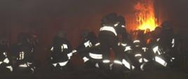Zdjęcia przedstawia strażaków ubranych w ubranie specjalne znajdujących się pomieszczeniu w którym jest pożar oraz silne zadymienie.
