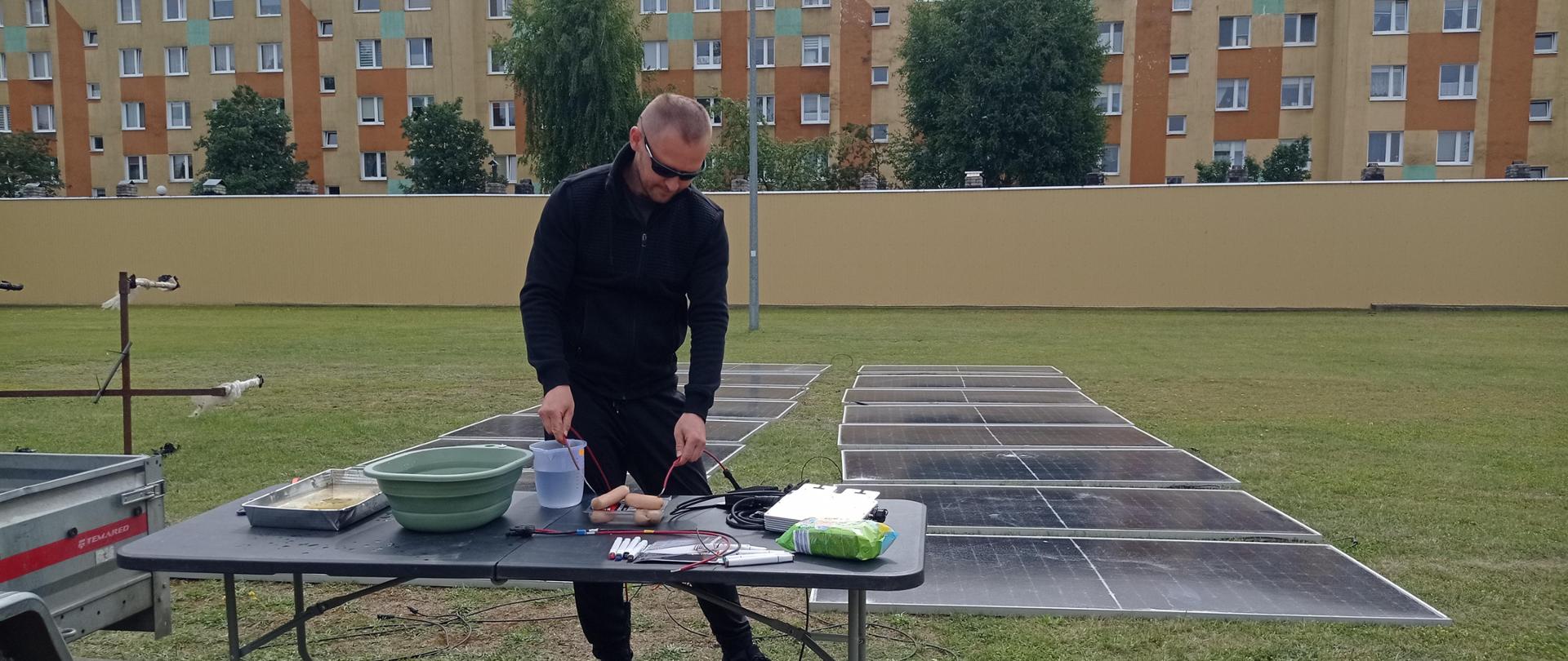 Na zdjęciu widać instruktora prowadzącego zajęcia. W sposób dosadny pokazuje skutki oddziaływania prądu stałego. Do pokazu wykorzystuje mięso wieprzowe a zgromadzeni wokół strażacy na własne oczy mogą zaobserwować jak powstaje łuk elektryczny oraz jak wielkim jest. zagrożeniem. Zajęcia prowadzone na placu wewnętrznym KP PSP w Wałczu. W tle na trawie rozłożona prowizoryczna instalacja fotowoltaiczna.