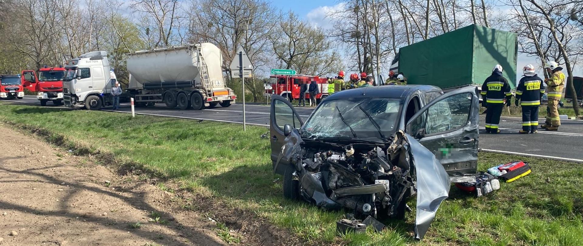 Zdjęcie przestawia samochody biorące udział w zdarzeniu jedn koloru ciemnego na poboczu drogi z rozbitym całkowicie przodem otwartymi drzwiami samochód typu ciężarowego z zielona plandeka na drugim poboczu,. przed samochodami stoi samochód ciężarowy z beczka który tarasuje całkowicie drogę. Przed samochodem ciężarowym stoją samochody strażackie . Pomiędzy samochodami strażacy.