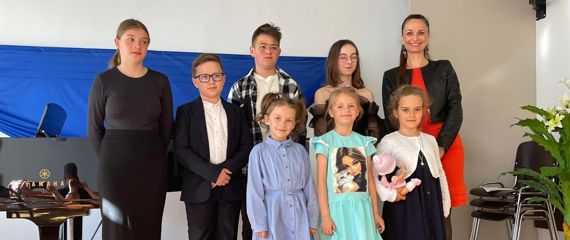 Na zdjęciu grupa uczniów: pięć dziewczynek i dwóch chłopców. Wszyscy wystąpili podczas koncertu klasy fortepianu. Obok nich stoi uśmiechnięta kobieta brunetka w czerwonej sukience i czarnej marynarce. Stoją na drewnianej podłodze. W tle za nimi na ścianie zawieszony jest niebieski materiał.