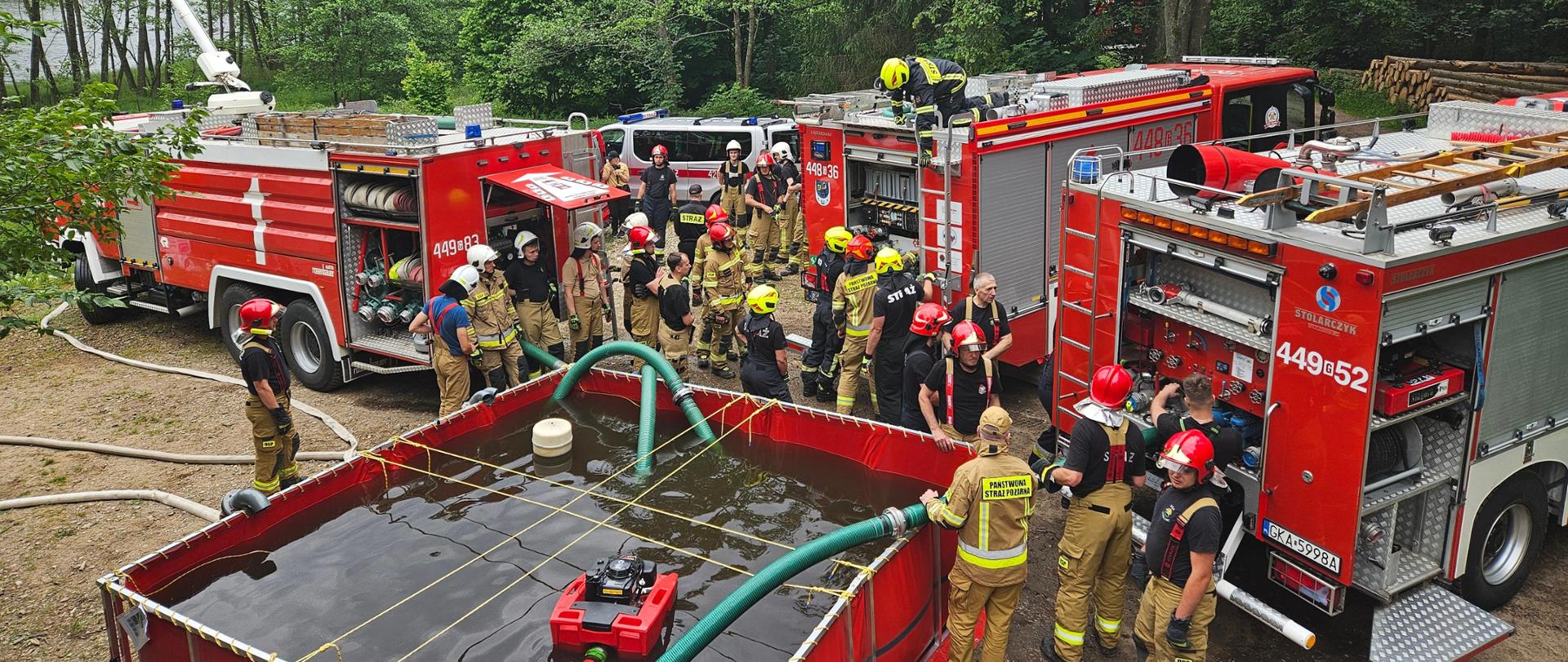 Zbiornik wody, przy którym ustawiono 3 samochody strażackie pomiędzy którymi znajdują się strażacy.