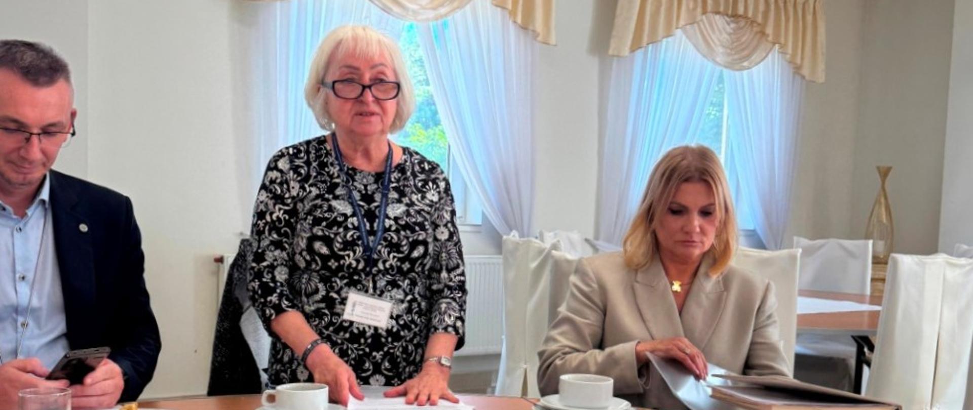 Spotkanie przy stole w jasnej sali z dużymi oknami i zasłonami; Kierownik WIORiN Oddziału w Płocku, pani Teresa Skóra stoi i mówi, obok niej siedzi mężczyzna korzystający z telefonu i kobieta przeglądająca dokumenty.