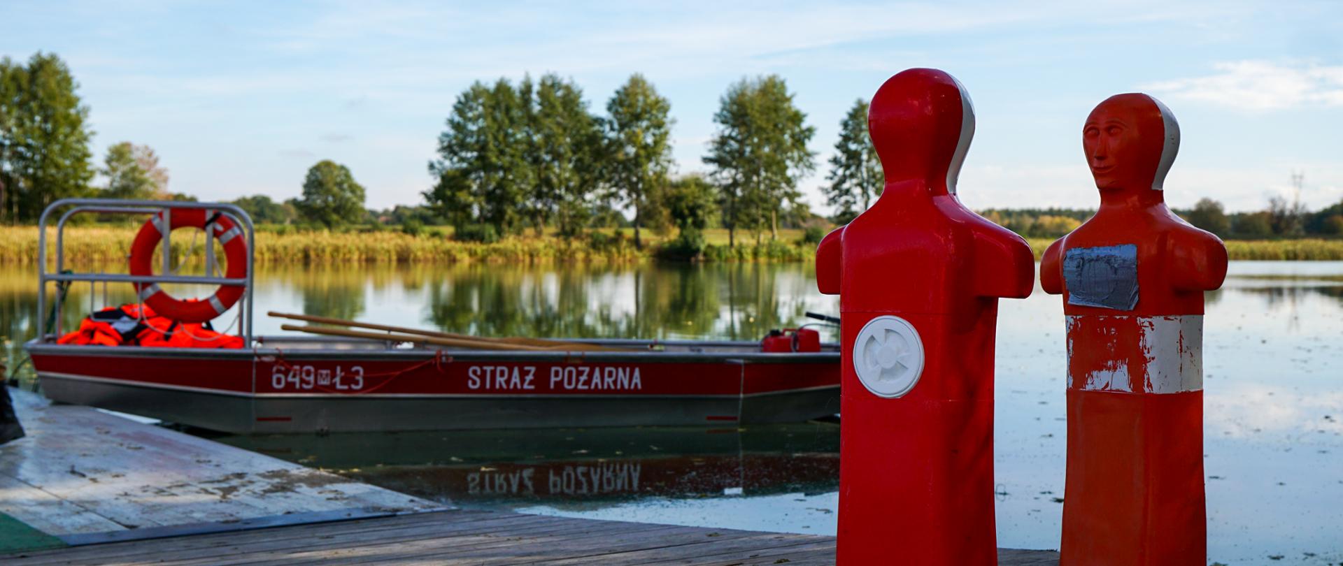 Fotografia przedstawia czerwoną łódź ratowniczą Państwowej Straży Pożarnej przytwierdzoną do drewnianego pomostu przy brzegu zbiornika wodnego. Po prawej stronie na pomoście stoją dwa jaskrawoczerwone fantomy medyczne w kształcie popiersi używane do ćwiczeń ratowniczych. Pogoda jest słoneczna, a na powierzchni spokojnej wody odbija się jasne niebo. W tle po drugiej stronie wody widoczne są zielone drzewa i pas roślinności. 