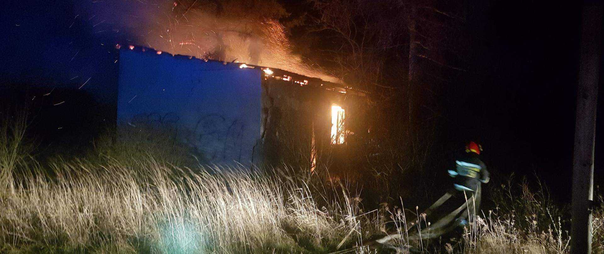 Strażak ubrany w sprzęt ochrony osobistej rozwija węże pożarnicze obok palącego się budynku. 