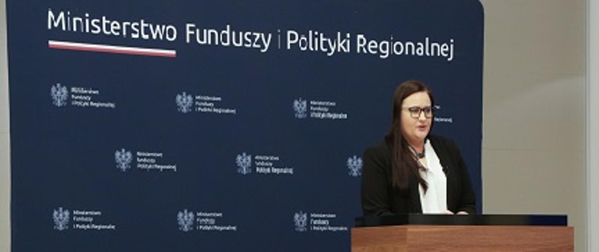 Minister Małgorzata Jarosińska-Jedynak przy mównicy, w tle ścianka z logo i nazwą Ministerstwa Funduszy i Polityki Regionalnej