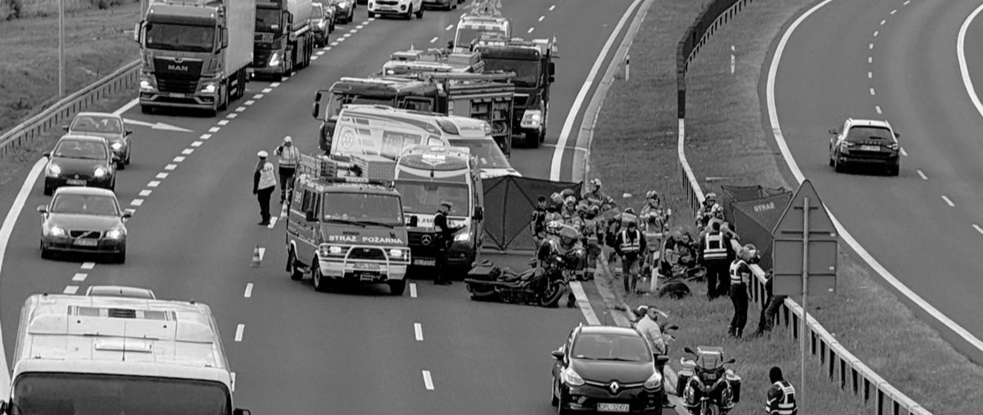 Czarno-białe zdjęcie przedstawia miejsce wypadku drogowego na wielopasmowej drodze szybkiego ruchu lub autostradzie.
Na pierwszym planie, po prawej stronie na pasie awaryjnym oraz na poboczu przy barierach ochronnych, zebrane są służby ratownicze:
* **Pojazdy służb:** Widać wóz strażacki oznaczony napisem „STRAŻ POŻARNA”, karetkę pogotowia ratunkowego oraz mniejsze pojazdy operacyjne i motocykle.
* **Miejsce zdarzenia:** Na pasie awaryjnym leży przewrócony motocykl. Obok rozstawiono parawany ochronne używane przez służby ratownicze. Wokół pracują ratownicy medyczni, strażacy oraz policjanci.
W tle widoczny jest duży zator drogowy:
* Ruch na pasach w kierunku zdarzenia został wstrzymany, tworząc długa kolejkę stojących samochodów osobowych i ciężarówek.
* Po prawej stronie, na przeciwległej jezdni oddzielonej pasem zieleni i barierami, ruch odbywa się płynnie.
* Krajobraz wokół drogi obejmuje wiadukt oraz teren leśny w tle.