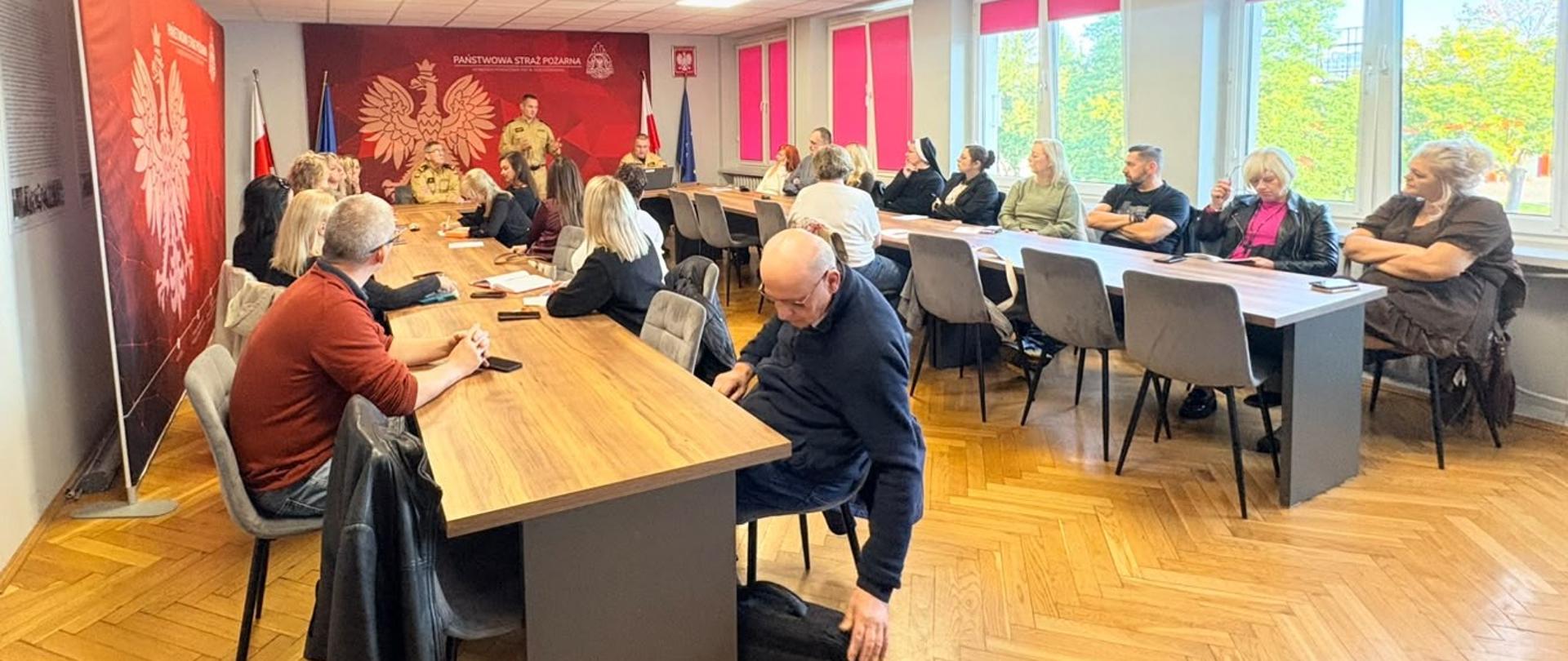 Spotkanie w sali konferencyjnej Państwowej Straży Pożarnej. Uczestnicy siedzą przy stołach w kształcie litery U i słuchają strażaka w mundurze znajdującego się na czele sali. Na czerwonej ścianie w tle widoczne jest godło Polski.
