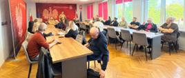 Spotkanie w sali konferencyjnej Państwowej Straży Pożarnej. Uczestnicy siedzą przy stołach w kształcie litery U i słuchają strażaka w mundurze znajdującego się na czele sali. Na czerwonej ścianie w tle widoczne jest godło Polski.