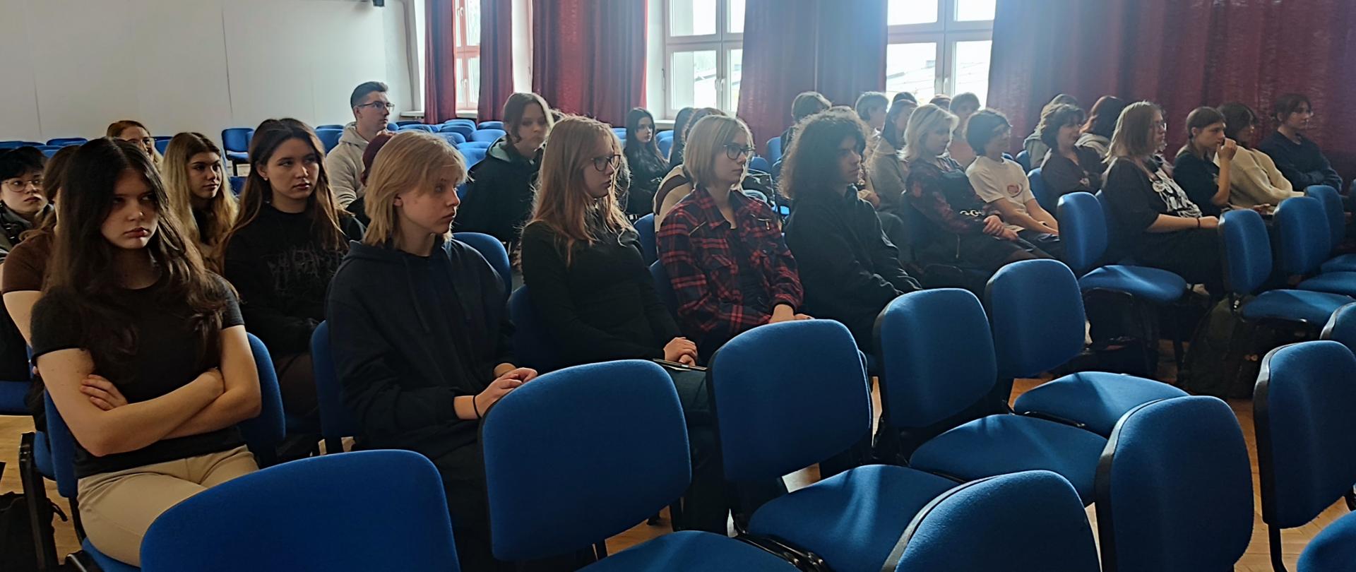 W auli szkolnej siedzą uczniowie na niebieskich krzesłach. Za nimi siedzi Paweł Gumiela. Przed nimi i za nimi jest dużo wolnych miejsc.