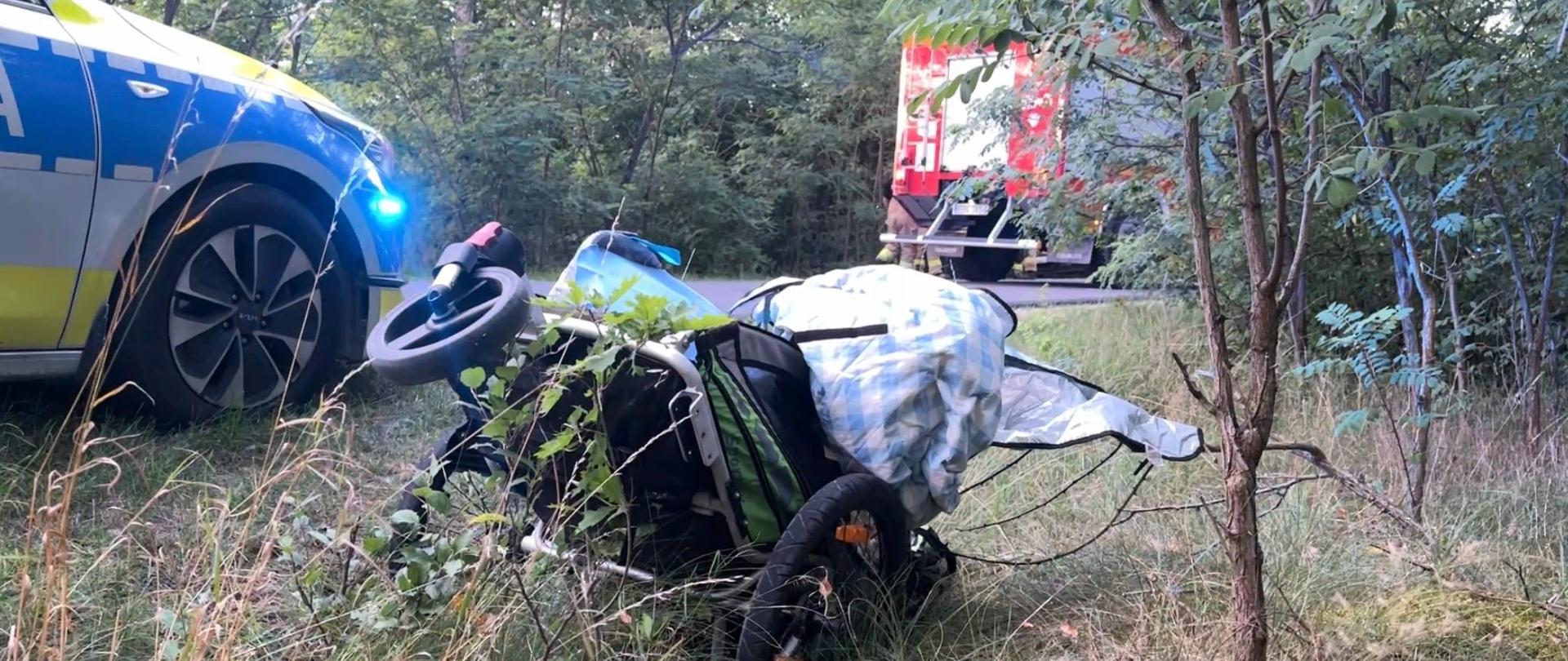 Na poboczu, wśród traw i krzaków, leży przewrócony wózek dziecięcy z dużymi kołami. Jest częściowo przykryty kocem lub materiałem w kratkę. Obok stoi radiowóz policyjny z włączonymi niebieskimi światłami, a w tle widać wóz strażacki.