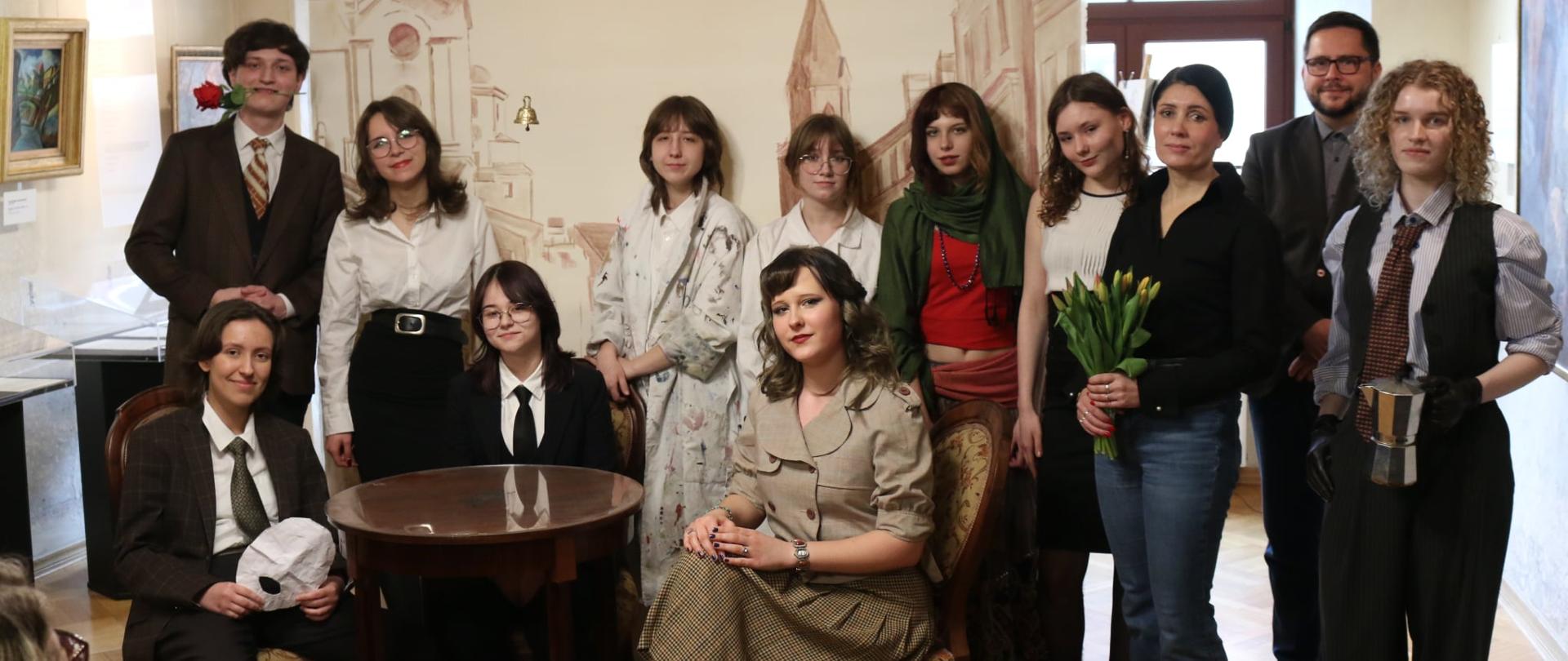 Grupa młodych ludzi w eleganckich ubraniach, część stoi, trzy osoby siedzą przy małym okrągłym stoliku. Wśród nich stoi kobieta z kwiatami, za nią mężczyzna w garniturze. Wszyscy patrzą w obiektyw. Za nimi mała ścianka z rysunkiem zabytkowych budowli. W przestrzeni wystawienniczej ze starymi obrazami