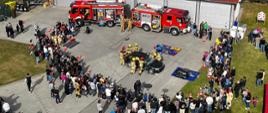 Tłum ludzi obserwuje strażaków w trakcie pokazu ratownictwa technicznego z użyciem dwóch wozów strażackich i rozcinanego samochodu na placu przed budynkiem PSP. Zdjęcie z lotu ptaka przedstawia publiczny pokaz strażacki zorganizowany na placu przed budynkiem z czerwonym dachem. Dwa wozy bojowe PSP stoją z otwartymi przedziałami sprzętowymi, wokół których zgromadził się tłum widzów, w tym dzieci i dorośli. W centralnej części placu kilku strażaków w ubraniach ochronnych i kaskach wykonuje działania ratownicze na rozbitym czarnym samochodzie, z wykorzystaniem narzędzi hydraulicznych. Teren jest zabezpieczony pachołkami i oznaczeniami. Po bokach widoczna jest duża grupa obserwatorów, część z nich to dzieci w kaskach lub przebraniach strażackich.
