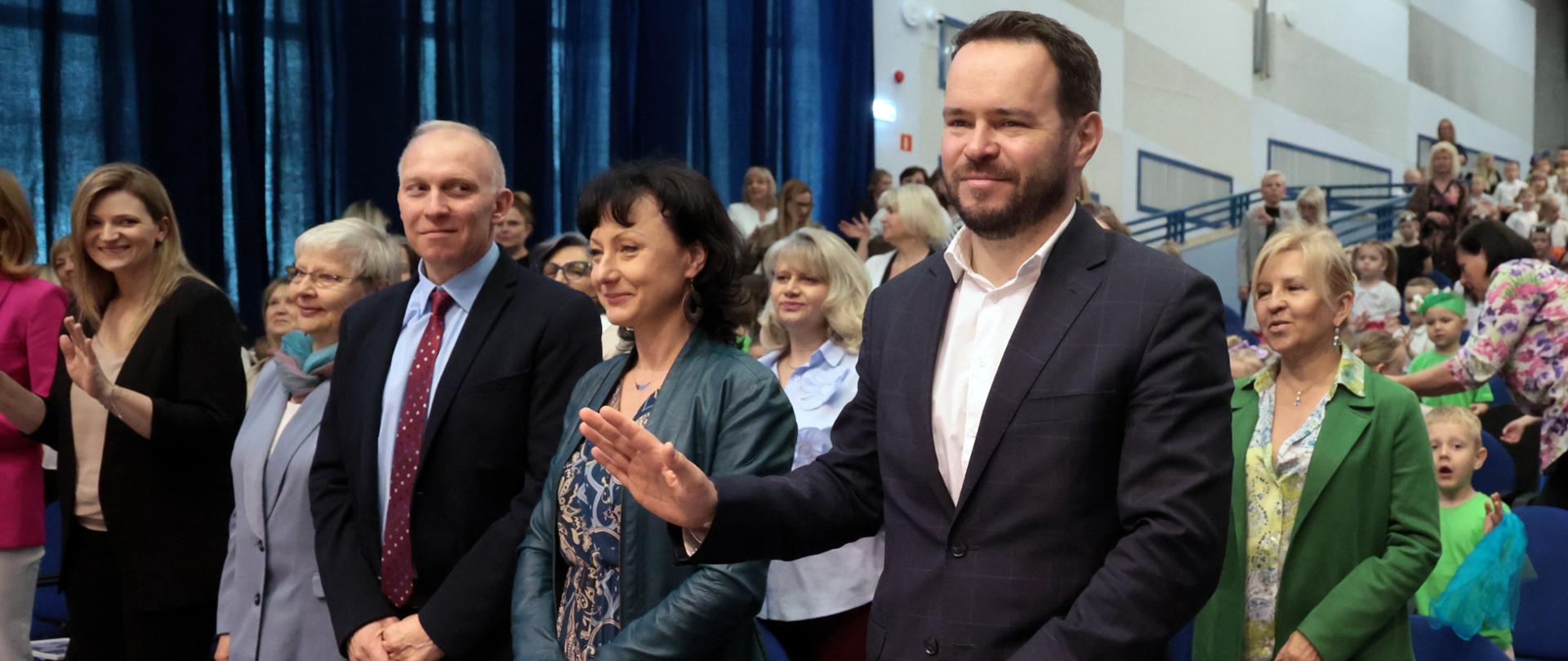 Kilka elegancko ubranych osób dorosłych stoi w pierwszym rzędzie widowni, trzymając oprawione podziękowania z napisem „Dziękujemy” i motywami kwiatowymi. W tle stoi publiczność, w której jest też kilkoro dzieci.