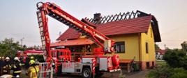 Na zdjęciu budynek mieszkalny po pożarze z widocznym spalonym dachem. Przed nim podnośnik hydrauliczny oraz strażacy prowadzący działania.