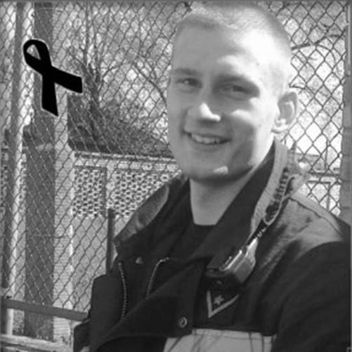 zdjęcie czarno białe młodego uśmiechniętego strażaka w ubraniu specjalnym z napisem straż. W tle widać płot
