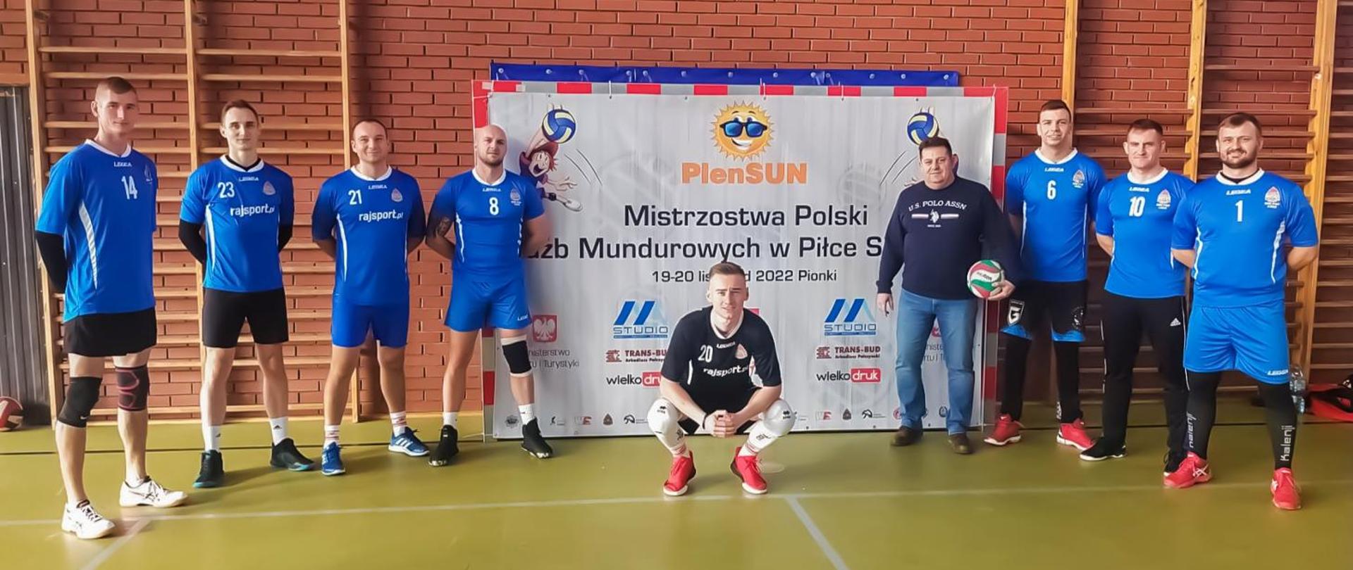 ośmiu siatkarzy oraz kierownik drużyny na tle ceglanej ściany, na ścianie znajdują się: drabinki gimnastyczne oraz baner Mistrzostwa Służb Mundurowych w Piłce Siatkowej"