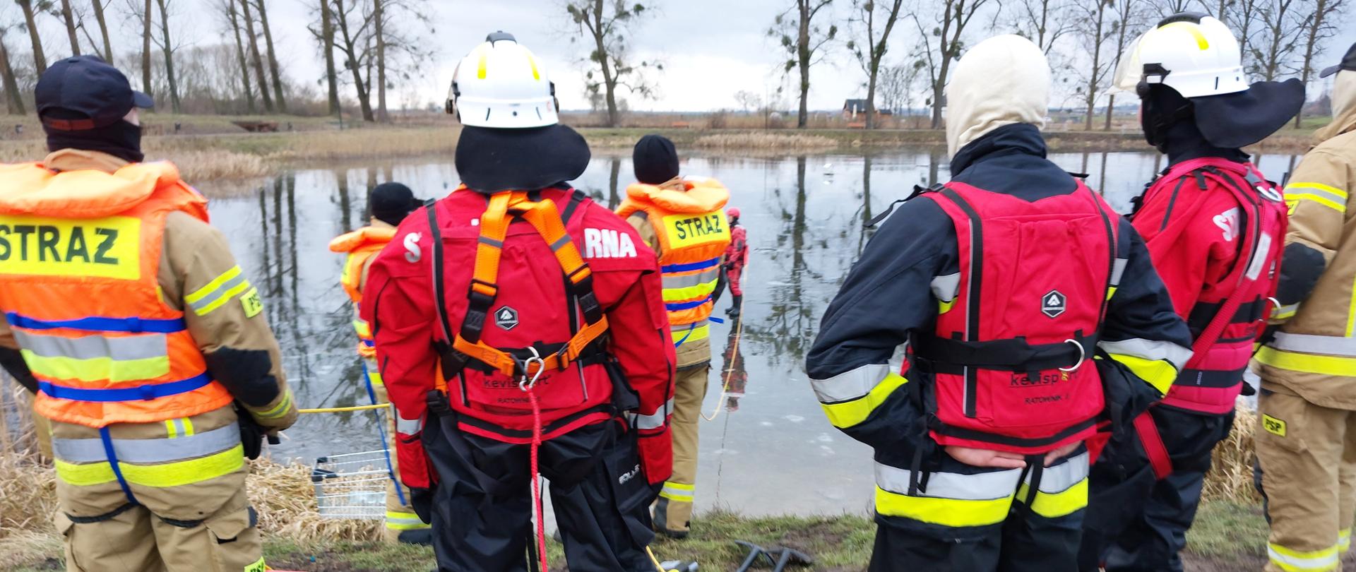 Strażacy i druhowie przyglądają się z brzegu przebiegowi ćwiczeń.