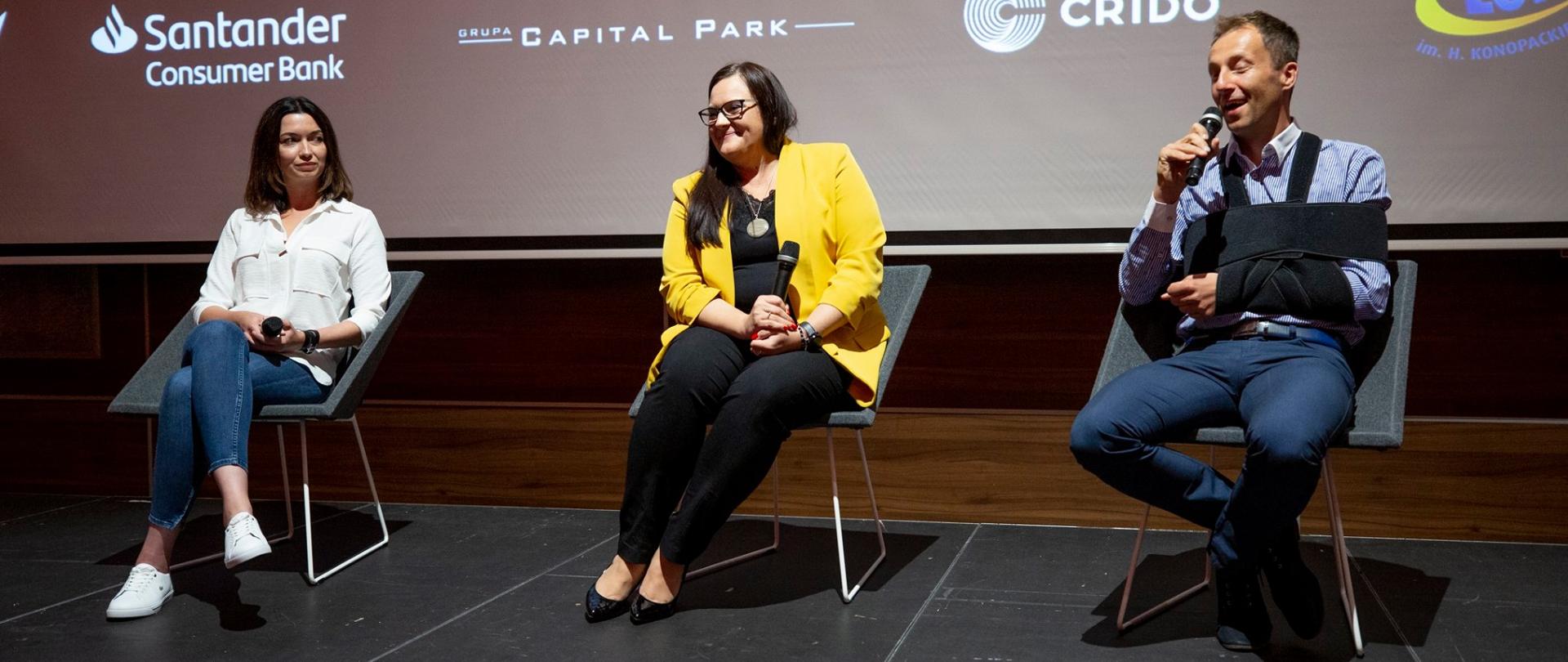 Na krzesłach siedzą trzy osoby. W środku minister M. Jarosińska-Jedynak.