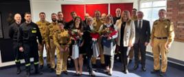 Grupa osób pozuje do wspólnego zdjęcia w pomieszczeniu z niebieską wykładziną i ceglaną ścianą. Wśród nich znajdują się funkcjonariusze Państwowej Straży Pożarnej w żółtych mundurach z polską flagą na ramieniu, strażacy w czarnych mundurach oraz cywilni pracownicy w eleganckich ubraniach. Trzy kobiety na pierwszym planie trzymają bukiety czerwonych róż i białych kwiatów, a jedna z nich dodatkowo elegancką torbę prezentową. W tle widoczne są czerwone tło z logo PSP oraz okno, przez które wpada naturalne światło. Grupa stoi blisko siebie, uśmiechając się do zdjęcia.