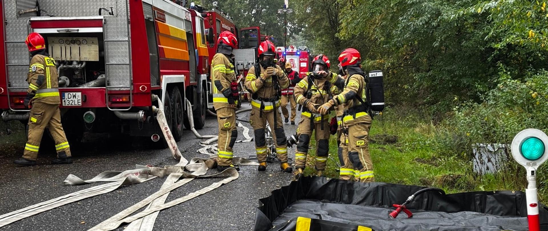 Strażacy PSP w aparatach ochrony dróg oddechowych przy samochodach gaśniczych; na jezdni rozłożone węże i czarny zbiornik przenośny.