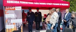 Namiot profilaktyczno-edukacyjny Komendy Powiatowej Państwowej Straży Pożarnej w Węgrowie, przygotowany dla uczestników uroczystości. Na pierwszym planie widoczne są materiały informacyjne, w tym baner kampanii „STOP pożarom traw!”. W tle stoją funkcjonariusze PSP i Policji w umundurowaniu wyjściowym oraz zaproszeni goście i mieszkańcy biorący udział w wydarzeniu.