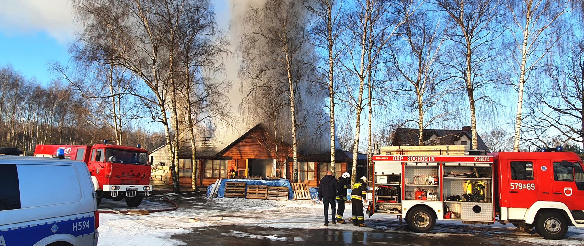Pożar budynku drewnianego jednokondygnacyjnego, pali się dach budynku, widoczny dym, który unosi się nad budynkiem, przed budynkiem dwa samochody pożarnicze oraz samochód policji.
