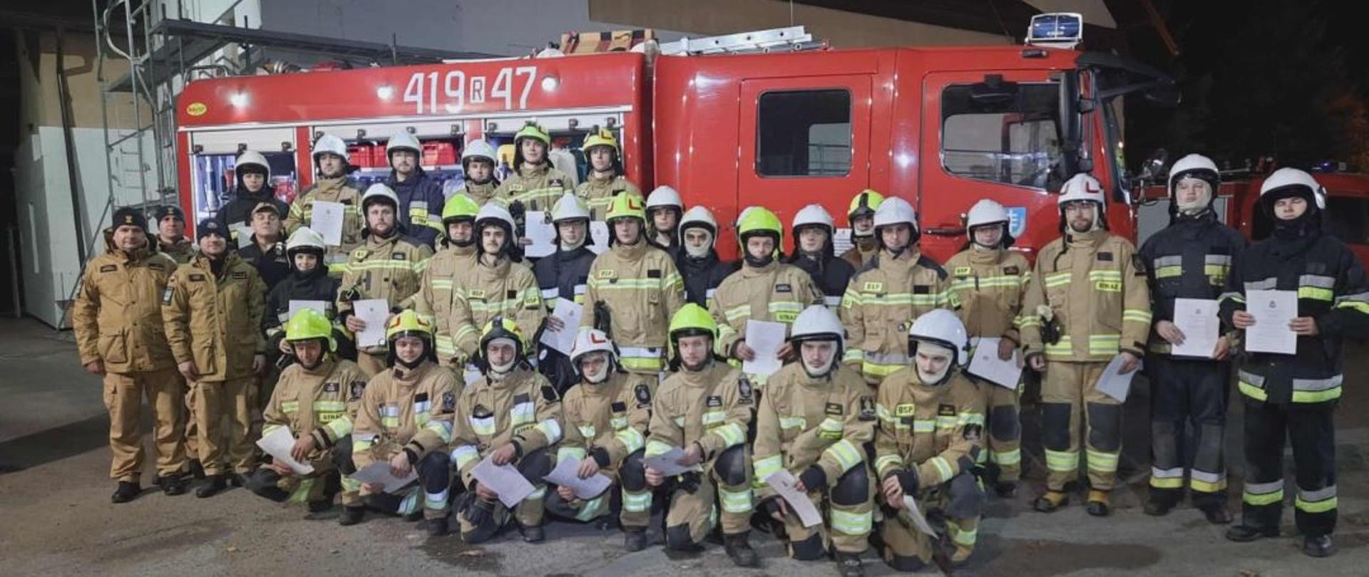 Kolorowa fotografia wykonana wieczorem przy sztucznym oświetleniu na placu remizy OSP Stara Wieś. Przestawia strażaków OSP w ubraniach specjalnych z hełmami trzymającymi zaświadczenia o ukończeniu szkolenia podstawowego OSP. Z boku w strojach służbowych stoją instruktorzy oraz Komendant Powiatowy PSP. W tle samochód pożarniczy.
