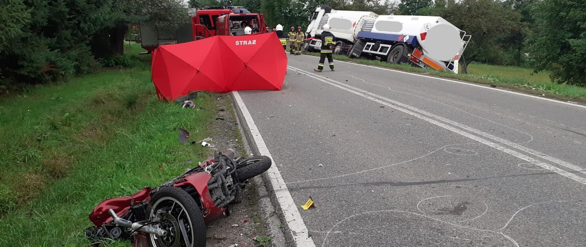 Zdjęcie obejmuje widok miejsca zdarzenia po wypadku. Po lewej stronie drogi wojewódzkiej u dołu zdjęcia leży rozbity motocykl. Patrząc w górę widać parawany strażackie zasłaniające osobę, która zginęła podczas zderzenia i samochody strażackie uczestniczące w akcji. Po prawej stronie jezdni widać uszkodzony samochód cysternę z przyczepą zabezpieczany wyciągarkami z pojazdów strażackich. W pobliżu samochodów znajduje się 7 strażaków PSP i OSP.