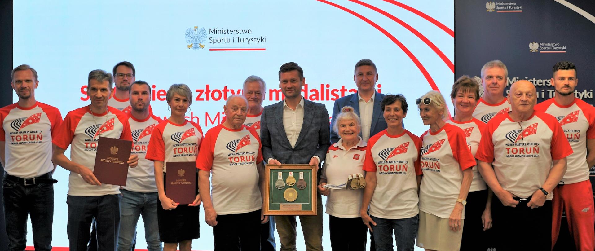 Minister Kamil Bortniczuk pozuje do zdjęcia ze złotymi medalistami Halowych Mistrzostw Świata w Lekkiej Atletyce Masters. Zawodnicy ubrani są w sportowe koszulki, a minister w garnitur. Minister trzyma w rękach podarunek od sportowców - medale halowych mistrzostw świata, które znajdują się za szybą w antyramie.