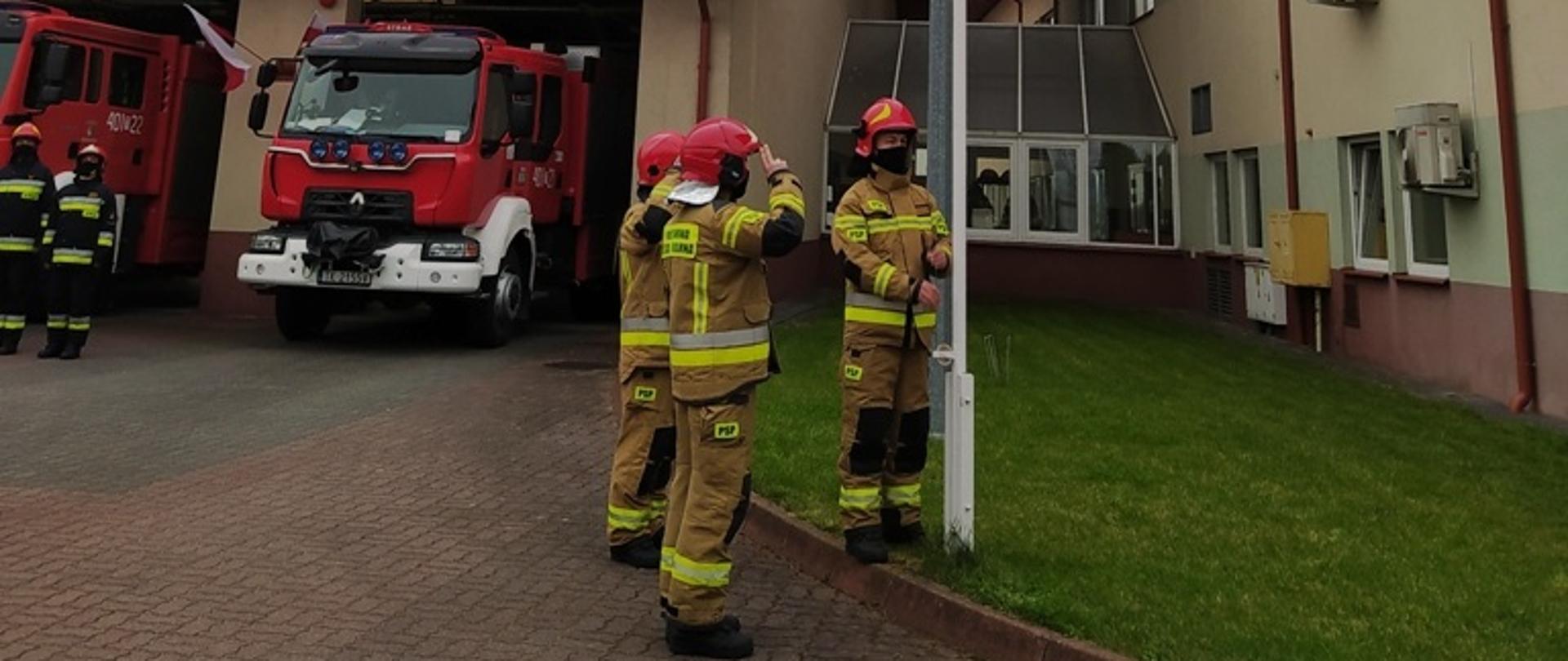 Zdjęcie przedstawia poczet flagowy przed masztem flagowym, gdzie jeden ze strażaków wciąga flagę na maszt, pozostali oddają honor. W tle pozostali strażacy ustawieni na uroczystej zbiórce przed pojazdami pożarniczymi 