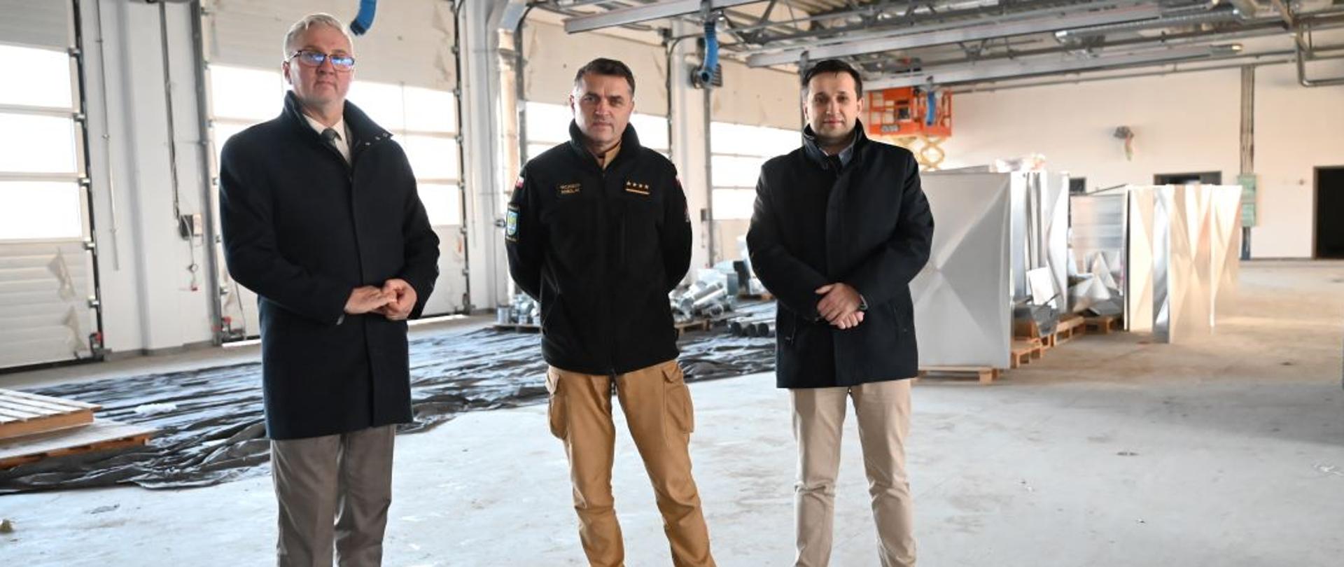 Kolorowa fotografia wykonana wewnątrz budowanej Komendy Powiatowej PSP w Brzozowie. Przedstawia strażaka w umundurowaniu służbowym stojącego między samorządowcami. Fotografia wykonana w części garażowej komendy.