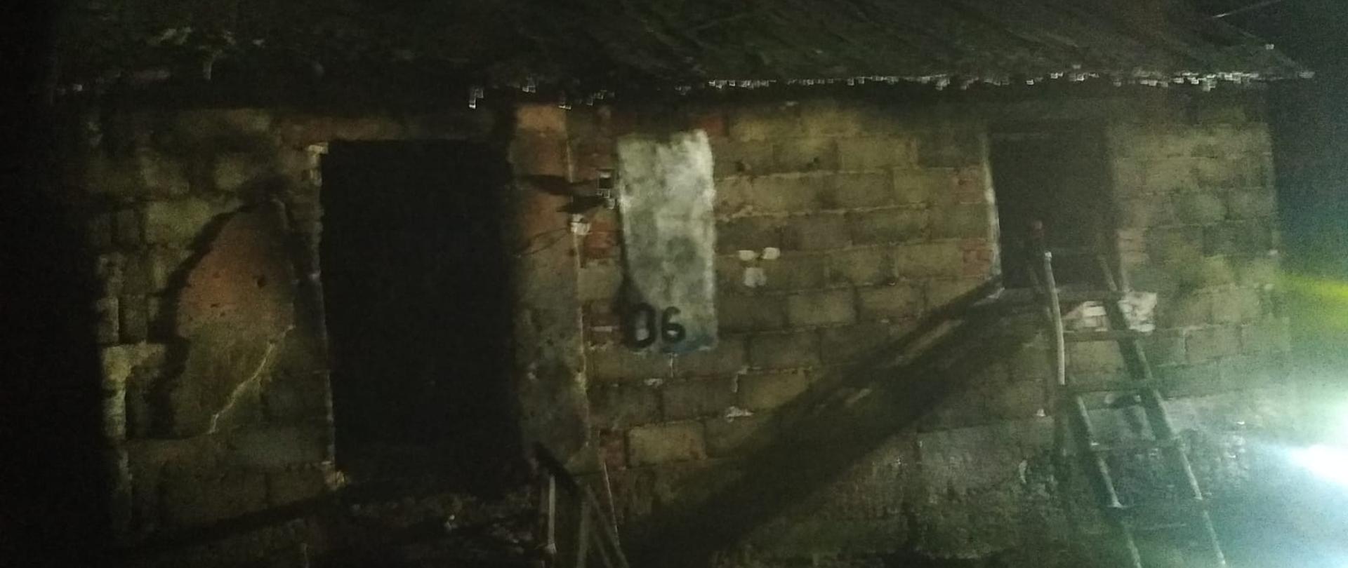 Zdjęcie przedstawia parterowy budynek z pustaków. Po lewej stronie zdjęcie otwarte drzwi, po prawej otwór po oknie do którego przystawiona jest drabina. Pora nocna.