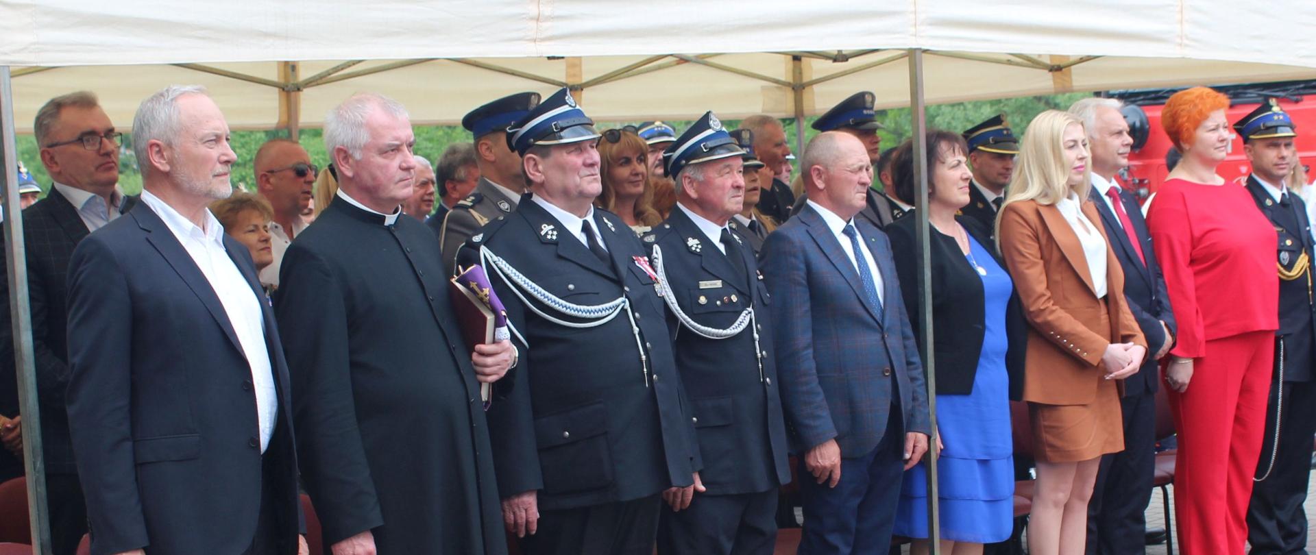 100-lecie jednostki Ochotniczej Straży Pożarnej w Chruszczewce (powiat sokołowski) strażacy w mundurach i goście stoją w szeregu pod namiotem, przed jednostką osp w tle czerwony samochód osp