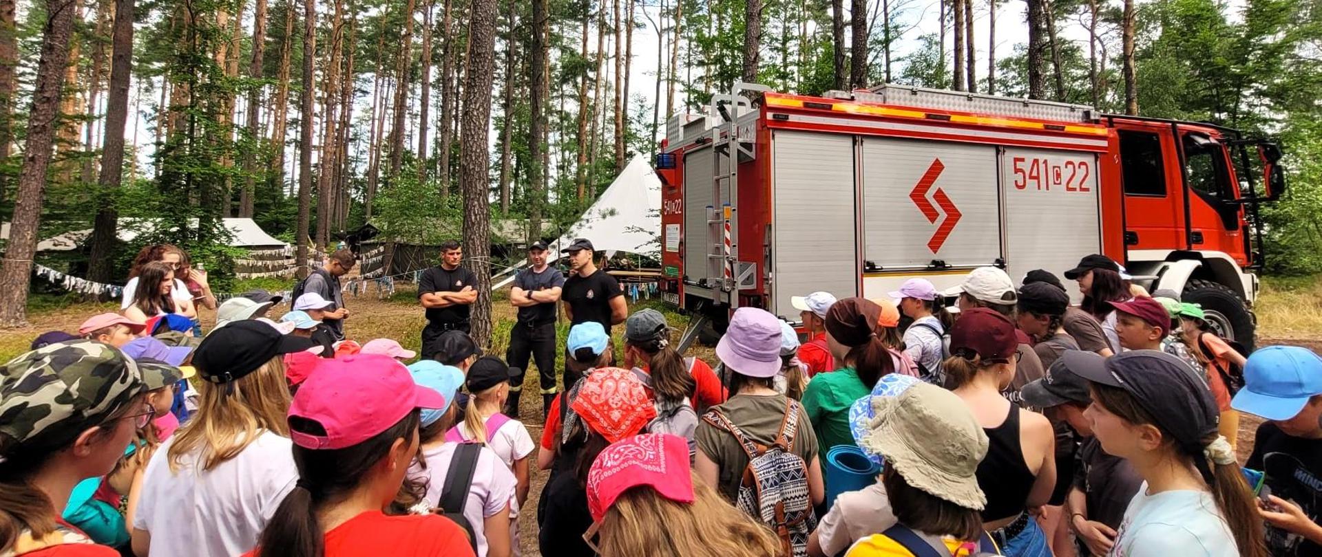 Trzech strażaków stoi naprzeciwko grupy dzieci. Strażacy są ubrani w czarne mundury. Po prawej stronie obok strażaków widać wóz strażacki. Wóz strażacki jest koloru czerwono-szaro-żółtego. Dzieci są ubrane na kolorowo. Część z dzieci ma na głowach kolorowe czapki i chustki. W tle widać wysokie drzewa. Za drzewami widać dachy budynków. Wszyscy znajdują się w lesie. 