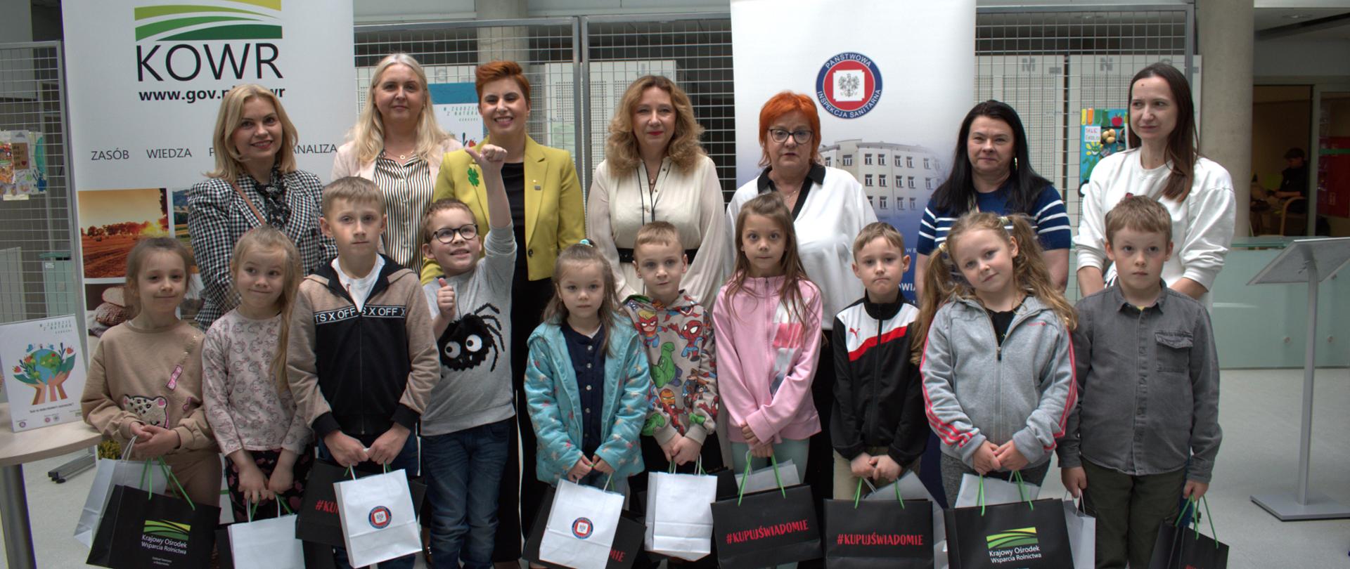 Na zdjęciu znajdują się laureaci I miejsca w konkursie „W zgodzie z naturą”. W tle znajdują się banery Krajowego Ośrodka Wsparcia Rolnictwa oraz Państwowej Inspekcji Sanitarnej. Od lewej na górze: członkowie jury oraz opiekunka grupy dzieci. Dzieci trzymają w rękach nagrody i są uśmiechnięte. Jeden chłopczyk macha ręką do kamery. 