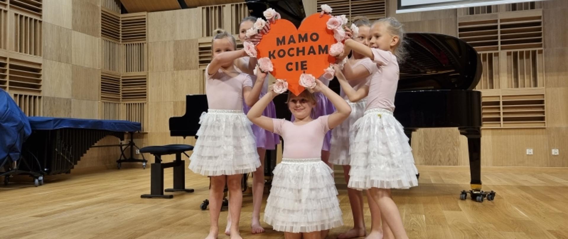 Dzieci w strojach baletowych z napisem " MAMO KOCHAM CIĘ" w kształcie serca.