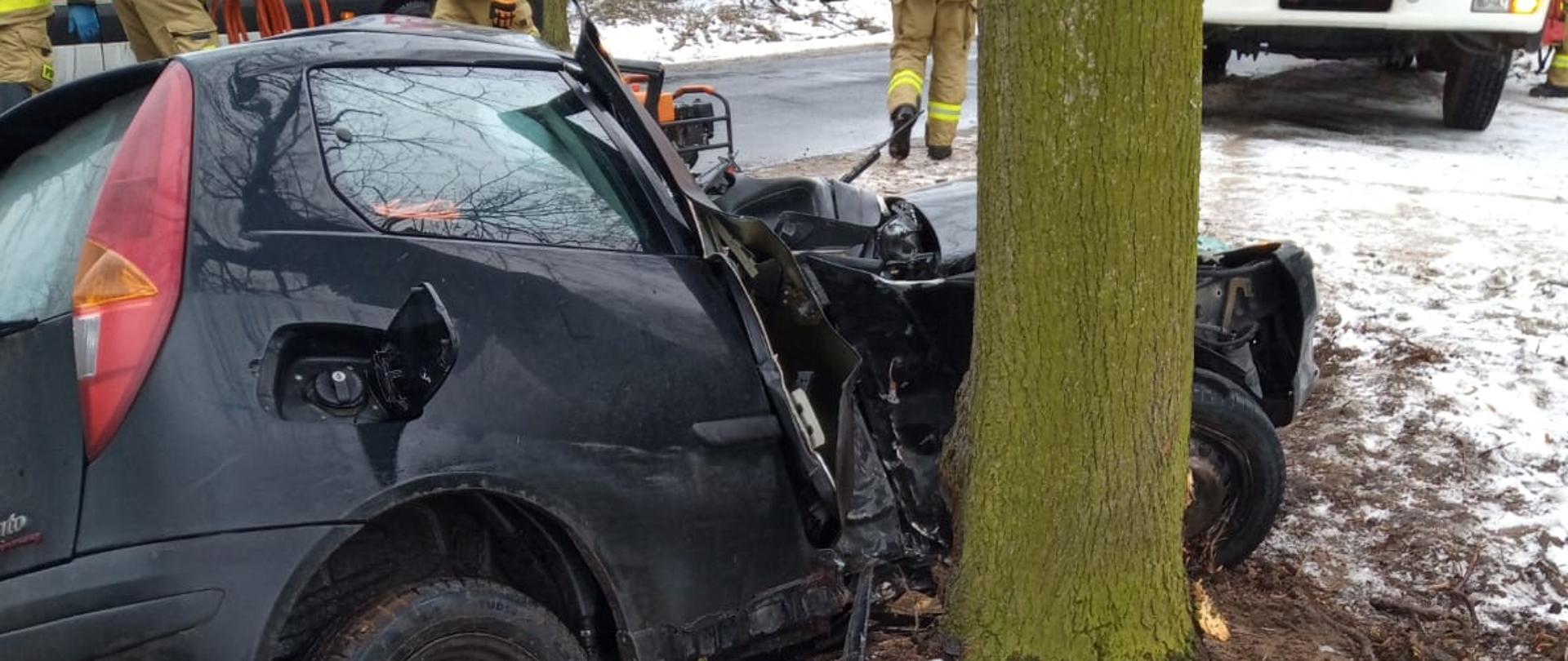 Zdjęcie prezentuje czarny samochód marki fiat punto, który uderzył w drzewo. Samochód jest przechylony, widać uszkodzoną prawą stronę samochodu. Uszkodzenia obejmują wgniecione drzwi pasażera oraz zgniecioną przednią część dachu. W tle widać 4 pracujących strażaków oraz karetkę pogotowia ratunkowego i samochód ochotniczej straży pożarnej. 