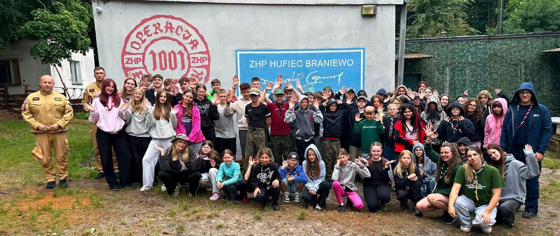Strażacy z grupą około sześćdziesięciu harcerzy na tle budynku w bazie harcerskiej.