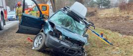 Zdjęcie przedstawia uszkodzony samochód znajdujący się w rowie drogowym. W tle widać samochody Państwowej Straży Pożarnej, Policji, Pomocy Drogowej oraz drzewa.