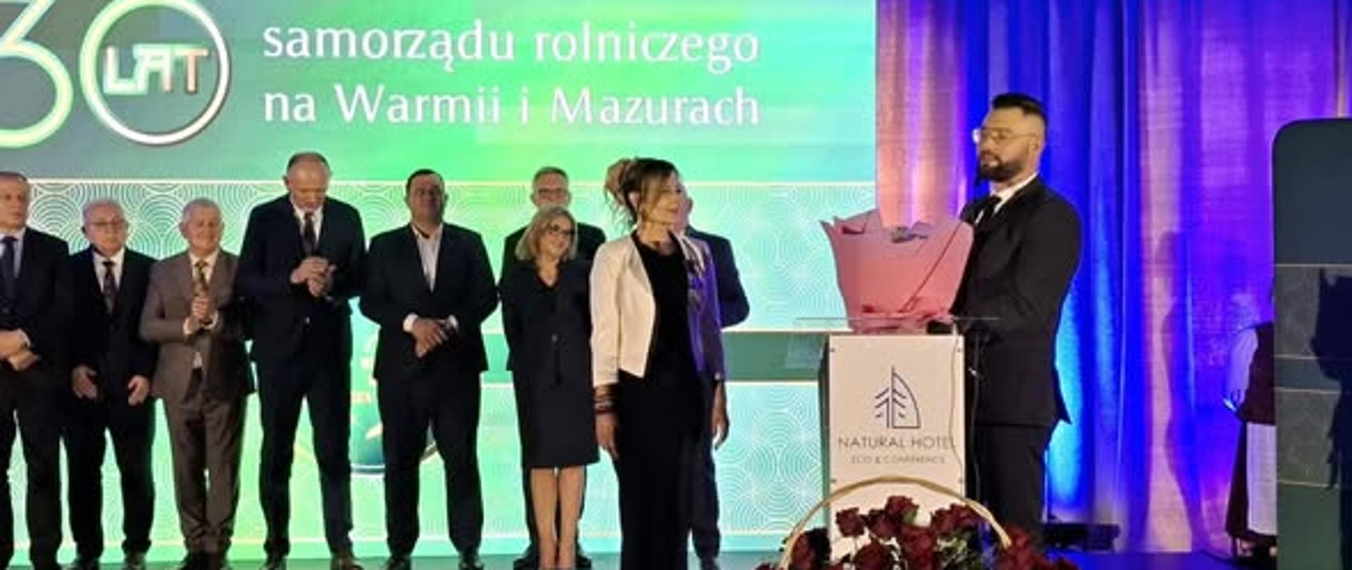 Na scenie po prawej stronie przed mównicą stoi mężczyzna, w głębi stoją równe osoby w strojach galowych, za ich plecami wyświetlany jest ekran 30 lat samorządu rolniczego na Warmii i Mazurach