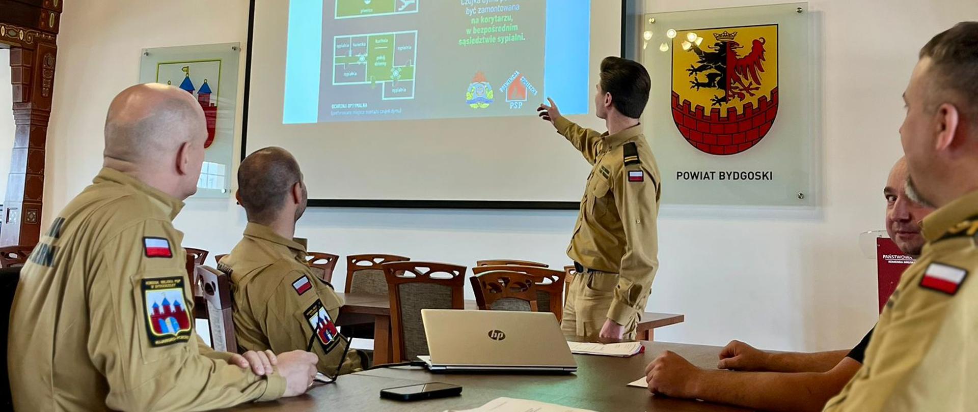 Na pierwszym planie stół, przy którym siedzi 4 strażaków. W tle jeden ze strażaków prowadzący zajęcia. 