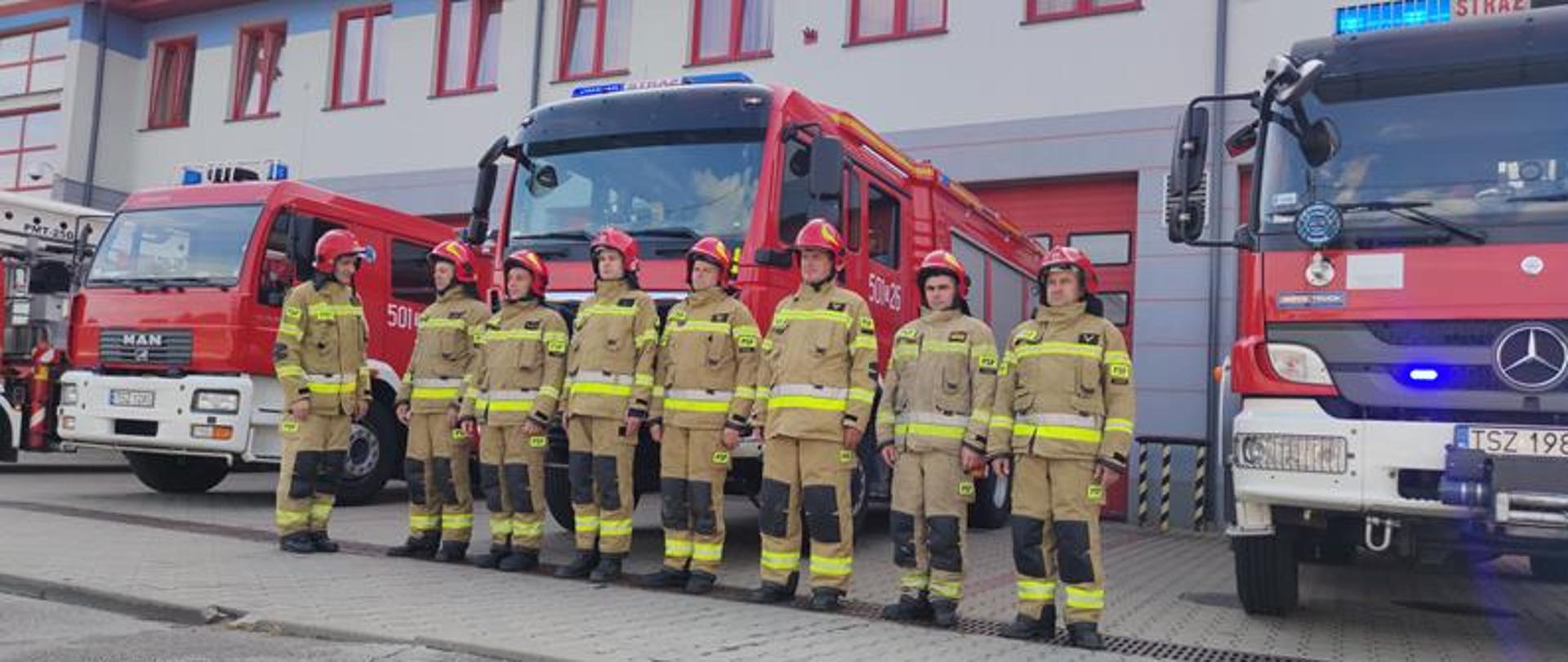 Strażacy ubrani w ubrania specjalne stoją przed samochodami oddając hołd poległym w powstaniu warszawskim.